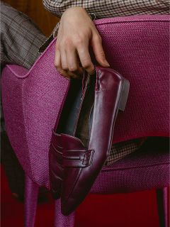 Una persona con pantalones a cuadros está sentada en una silla rosa con una superficie texturizada, sosteniendo un mocasín burdeos.