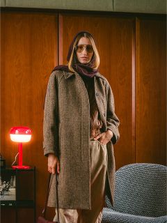 Una persona con un abrigo marrón y una bufanda está de pie en una habitación junto a una lámpara roja y una silla gris, con paredes con paneles de madera al fondo.