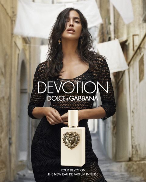 Una mujer vestida de negro se encuentra en un callejón estrecho detrás de una botella de perfume "Dolce & Gabbana Devotion". Al fondo, cuelga ropa blanca.