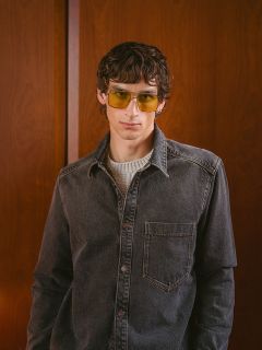 Un hombre con cabello ondulado, que lleva gafas de sol amarillas, una camisa vaquera gris y un suéter claro, está de pie frente a una pared de madera y mira a la cámara.