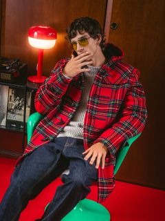 Un hombre con un abrigo de cuadros rojo y gafas de sol está sentado en una silla verde junto a una lámpara roja y sostiene su mano cerca de su cara en una pose elegante.