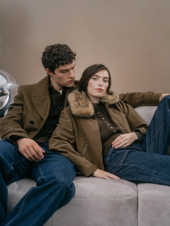 Un hombre y una mujer con chaquetas de color oliva están sentados juntos en un sofá claro.