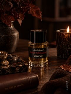 Sobre una mesa de madera, bajo una luz cálida y tenue, una botella del perfume "Musc Ravageur" se encuentra junto a una vela encendida, libros y hojas de otoño.