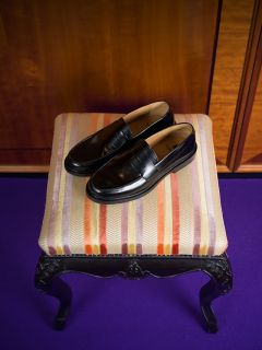 Un par de mocasines de cuero negro se encuentran sobre un taburete acolchado a rayas con patas negras ornamentadas, que descansa sobre una alfombra morada.