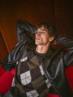 Una persona con cabello rizado, que lleva una chaqueta de cuero y un suéter de argyle, se relaja en un sillón rojo, recostada y con las manos entrelazadas detrás de la cabeza.