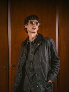 Una persona con gafas de sol y chaqueta oscura está de pie frente a puertas de madera, mirando ligeramente hacia un lado y con las manos en los bolsillos.