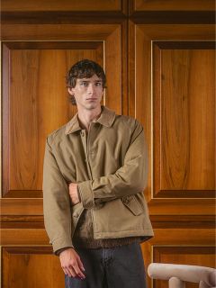 Un hombre con una chaqueta marrón claro está de pie frente a una puerta con paneles de madera y mira a la cámara con una expresión inexpresiva.