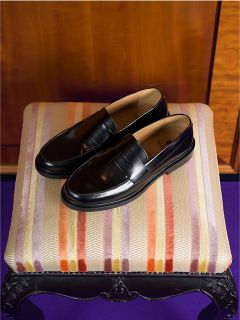 Un par de mocasines de cuero negro están sobre un taburete acolchado a rayas, detrás del cual hay una pared de madera.