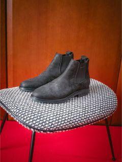 Un par de botas Chelsea negras en una silla con estampado blanco y negro frente a una alfombra roja y una pared de madera.