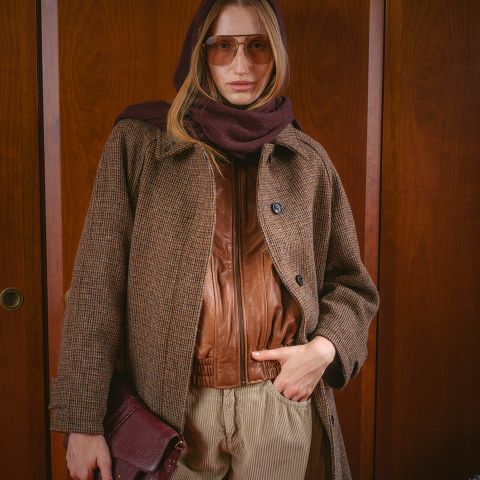 Una mujer con gafas de sol, una bufanda marrón, un abrigo y una chaqueta de cuero, sosteniendo un bolso y de pie frente a un fondo de madera.