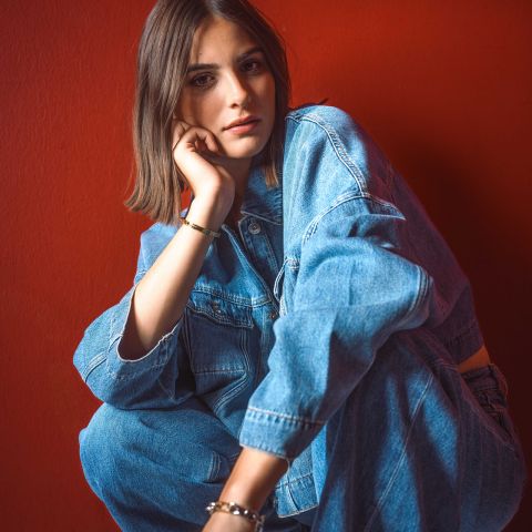 Una mujer con chaqueta vaquera y jeans está sentada frente a una pared roja brillante, apoyando la barbilla en la mano y mirando a la cámara.