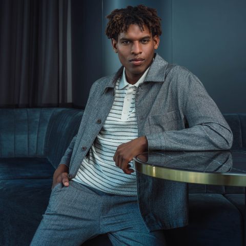 Un hombre con un traje gris y una camisa a rayas está sentado en un sofá azul, apoyando un brazo en una mesa de cristal y mirando a la cámara con confianza.