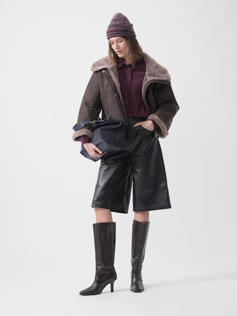 Una mujer con gorro, chaqueta de piel de cordero, camisa burdeos, culottes de cuero y botas de tacón, sosteniendo un bolso azul marino y de pie frente a un fondo blanco.