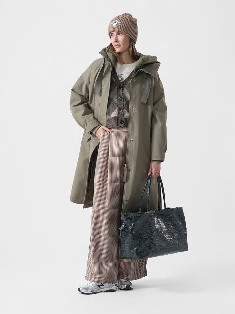 Una mujer con un abrigo verde, pantalones estampados, zapatillas y un gorro sostiene un bolso grande y texturizado, de pie frente a un fondo blanco liso.
