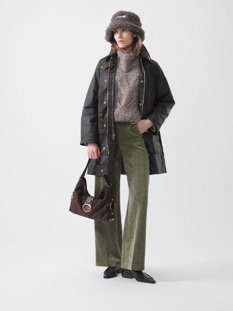 Una mujer con un gorro de piel, una chaqueta oscura, un suéter marrón, pantalones verdes de pana y zapatos negros, sosteniendo un bolso marrón y de pie frente a un fondo liso.