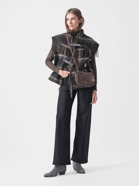 Una mujer con un poncho a cuadros, una blusa marrón, pantalones negros y botas grises, llevando un bolso bandolera marrón, posa frente a un fondo blanco.
