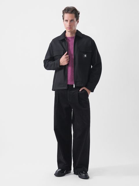 Un hombre posa con una chaqueta negra sobre un suéter morado, pantalones negros de pernera ancha y zapatos negros, frente a un fondo blanco liso.