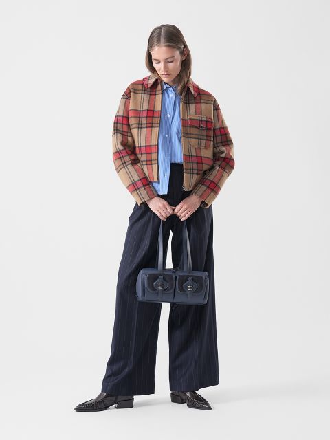Una mujer con una chaqueta a cuadros, una camisa azul y pantalones de rayas anchas sostiene un bolso de mano azul marino de dos compartimentos y se para frente a un fondo blanco liso.