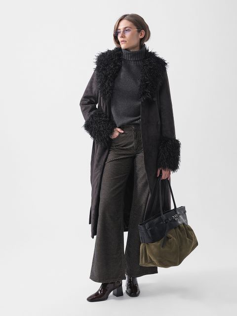 Una mujer está de pie, vestida con un abrigo oscuro ribeteado de piel, un jersey de cuello alto, pantalones acampanados y botas. Lleva un bolso bicolor y gafas, y posa con confianza.
