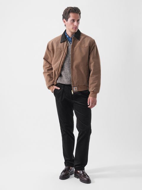 Un hombre que lleva una chaqueta marrón, un suéter gris, una camisa azul, pantalones negros y zapatos oscuros, de pie frente a un fondo blanco liso.