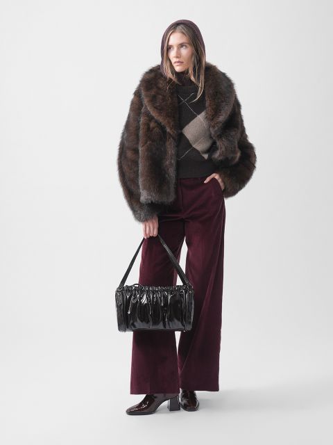 Una persona con un abrigo de piel, un suéter estampado, pantalones anchos color borgoña y botas sostiene un bolso de mano negro brillante y se para frente a un fondo blanco liso.