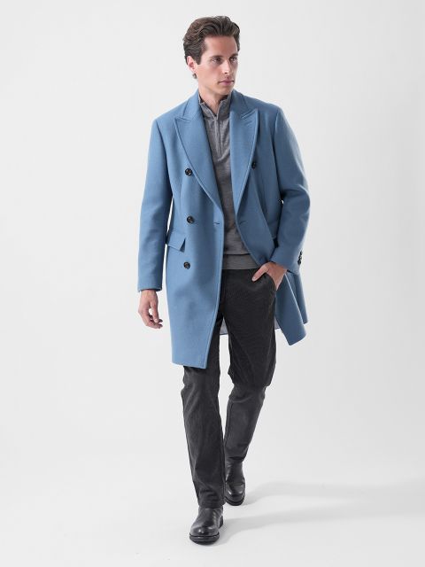 Un hombre con un traje cruzado azul claro, una camisa gris, pantalones oscuros y botas negras, caminando con confianza frente a un fondo blanco liso.
