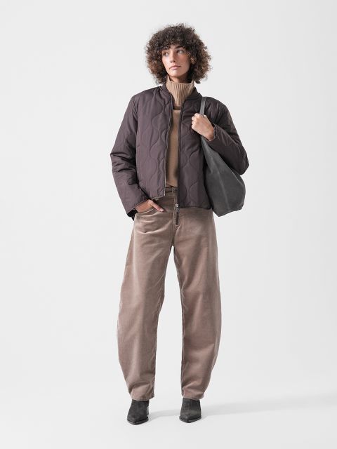 Una persona con una chaqueta acolchada marrón, un jersey de cuello alto marrón claro, pantalones anchos beige y zapatos negros, sosteniendo una bolsa de mano negra y de pie frente a un fondo liso.