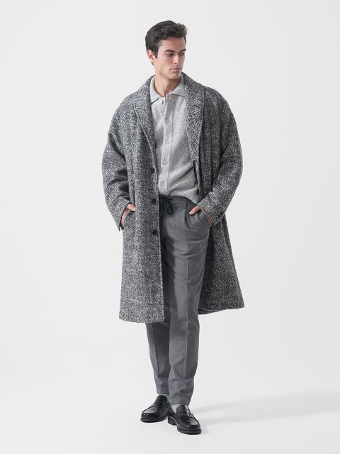 Un hombre que lleva un abrigo gris texturizado, una camisa gris claro con botones, pantalones grises y zapatos negros, de pie con las manos en los bolsillos frente a un fondo blanco.