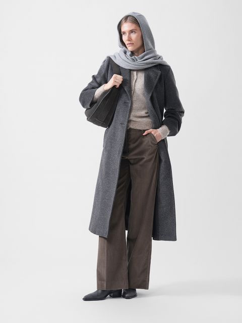 Una mujer con un abrigo gris con capucha, un suéter beige, pantalones anchos marrones y zapatos negros, sosteniendo un bolso negro y de pie frente a un fondo blanco.