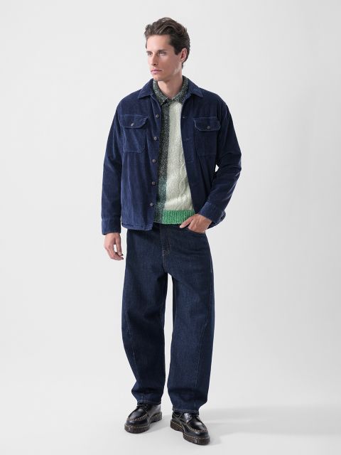 Un hombre con una chaqueta azul marino, un suéter verde y blanco, jeans oscuros holgados y zapatos negros, de pie frente a un fondo blanco liso.