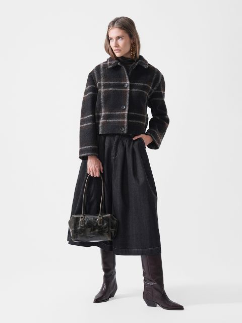 Una mujer con una chaqueta a cuadros, una falda negra y botas altas marrones, sosteniendo un bolso de mano negro, de pie frente a un fondo blanco liso.