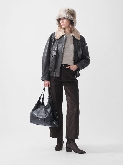 Una mujer con un gorro de piel, una chaqueta de cuero negra, un suéter beige, pantalones marrones y botas de tacón, sosteniendo un bolso negro grande y de pie frente a un fondo blanco.