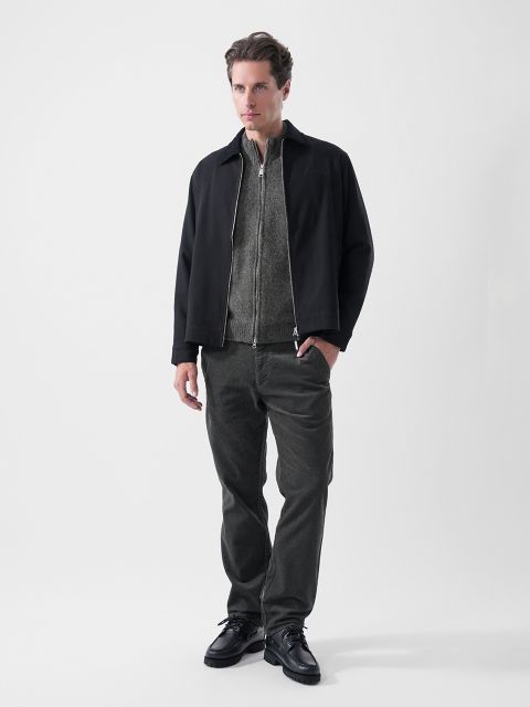 Un hombre de pie frente a un fondo de color liso, con una chaqueta negra, un jersey gris con cremallera, pantalones oscuros y zapatos negros, mirando ligeramente hacia un lado.
