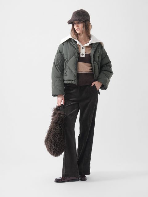 Una mujer con una chaqueta de plumas verde, un suéter a rayas, pantalones marrones y un gorro de piel, sosteniendo un bolso marrón esponjoso y de pie frente a un fondo blanco liso.