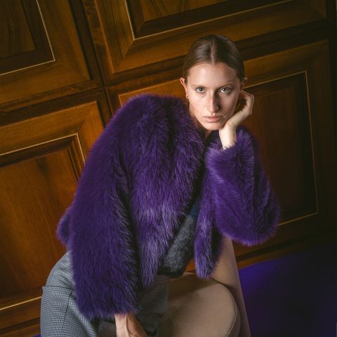 Una persona con un abrigo de piel sintética morado se apoya en una silla, mirando a la cámara, con paredes con paneles de madera al fondo.