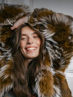 Una mujer sonriente con un abrigo de piel sintética marrón y blanco de pelo largo, con las manos entrelazadas detrás de la cabeza.