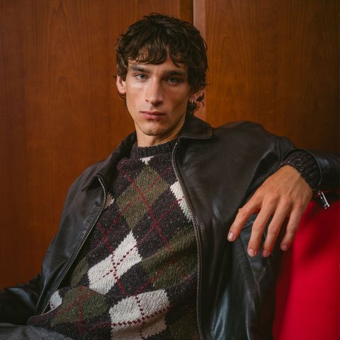 Un joven con una chaqueta de cuero oscura y un suéter de argyle está sentado en una silla roja, posando frente a una pared de madera y mirando a la cámara.