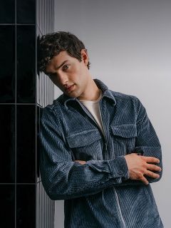 Un hombre con una camisa de pana azul con cremallera, con los brazos cruzados y apoyado en una pared de azulejos oscuros.
