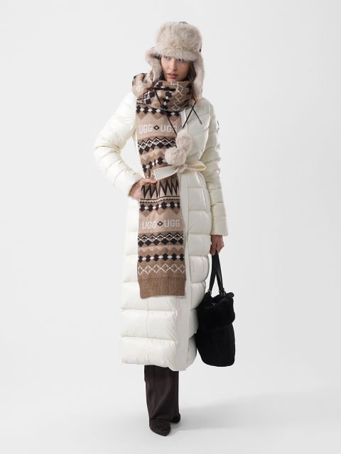 Una mujer lleva un largo abrigo de plumas blanco, una bufanda con estampado marrón con el logo de UGG y un gorro de trampero de piel sintética; en la mano tiene un bolso de mano negro.