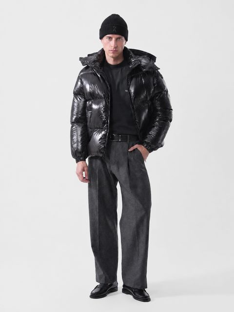 Un hombre con gorra negra, chaqueta acolchada negra brillante, camisa negra, pantalones grises y mocasines negros.