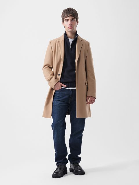 Un hombre que lleva un abrigo color canela, un suéter azul marino con cremallera en el cuello, una camiseta blanca, jeans azul oscuro y zapatos negros.