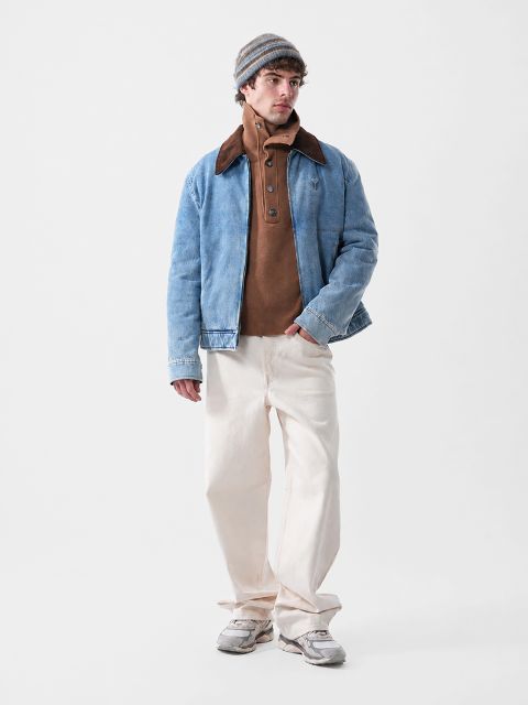 Un hombre con una chaqueta de mezclilla azul sobre un suéter marrón abotonado, pantalones color crema y un gorro.