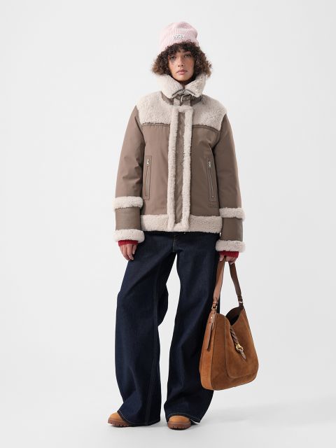 Una modelo con chaqueta de color marrón claro con ribete de piel sintética color crema, jeans anchos azules y gorro rosa, sosteniendo un bolso de hombro de gamuza marrón.