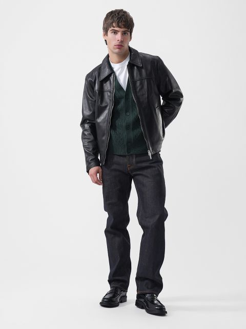 Un hombre que lleva una chaqueta de cuero negra sobre un cárdigan verde, una camiseta blanca, vaqueros oscuros y mocasines negros.