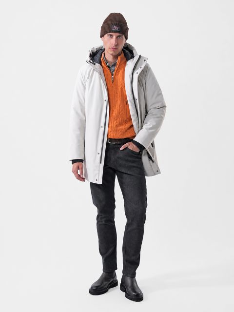Un hombre que lleva un parka blanco con capucha, un suéter naranja con cremallera de un cuarto, pantalones vaqueros negros y un gorro marrón.