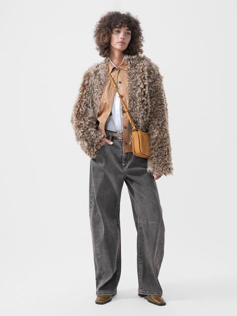Una persona que lleva una chaqueta con textura esponjosa sobre una camisa marrón claro con cuello blanco, combinada con unos vaqueros grises de pernera ancha y un bolso bandolera marrón claro.