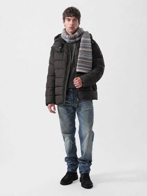 Un hombre que lleva una chaqueta acolchada de color marrón oscuro, un suéter gris, una bufanda a rayas, vaqueros azules y zapatos oscuros.