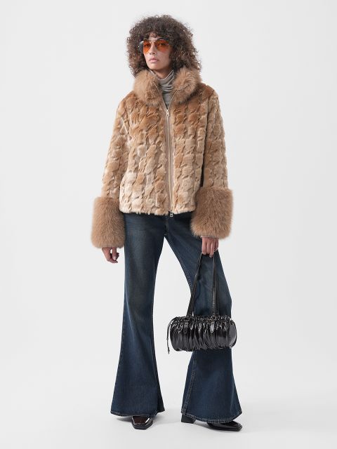 Una persona con una chaqueta de piel sintética de color tostado con puños anchos, vaqueros acampanados oscuros y gafas de sol naranjas, sosteniendo un bolso de mano negro plisado.