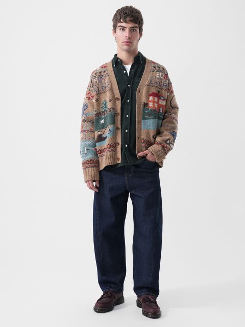 Un hombre con un cárdigan marrón claro estampado, una camisa abotonada verde oscuro y unos vaqueros azul oscuro.