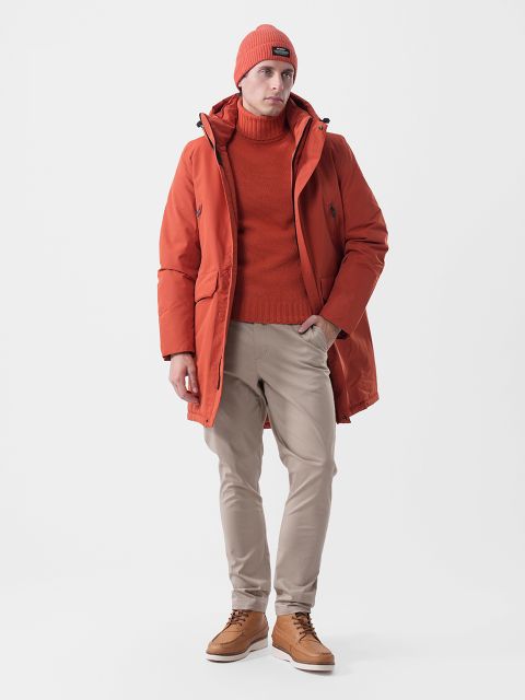 Un hombre con un gorro naranja, un jersey de cuello alto y una parka, así como pantalones beige y botas de cuero marrones.